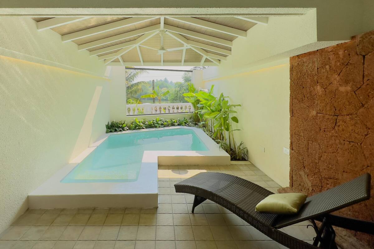 A pool area with a ceiling structure, a lounge chair, and potted plants.