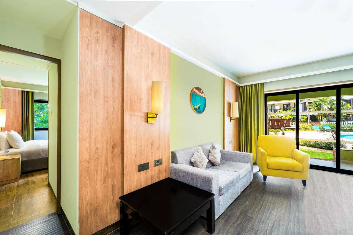 A hotel room with a bed, a sofa, a yellow armchair, a coffee table, and a view of a pool and greenery.