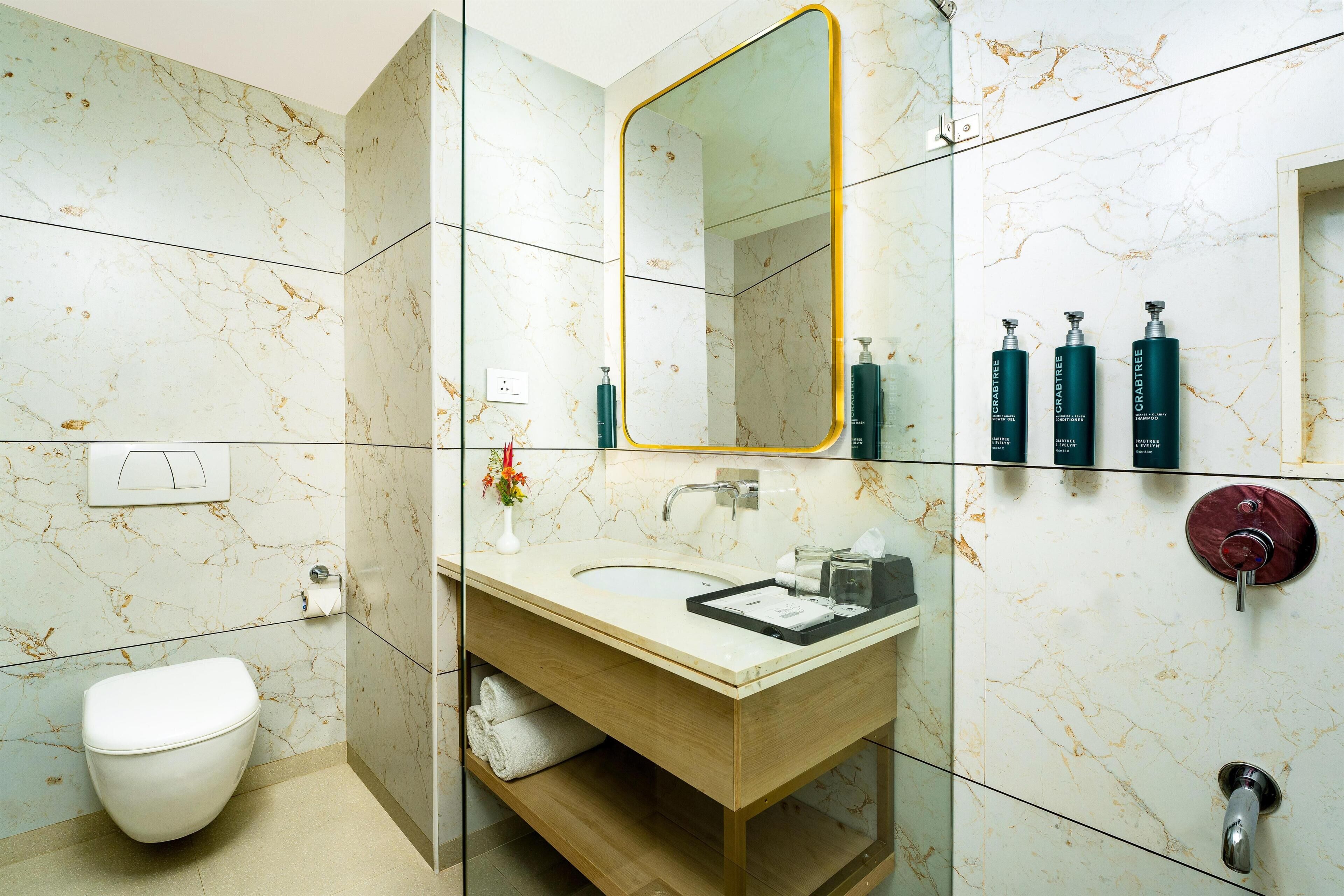 Een badkamer met een marmeren wand, een houten wastafelmeubel, een wastafel, een toilet en een spiegel met gouden lijst.