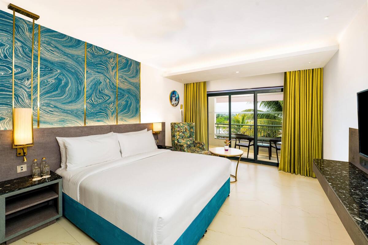 A hotel room with a large bed, a desk with a lamp, a chair, a TV, and a balcony with yellow curtains.