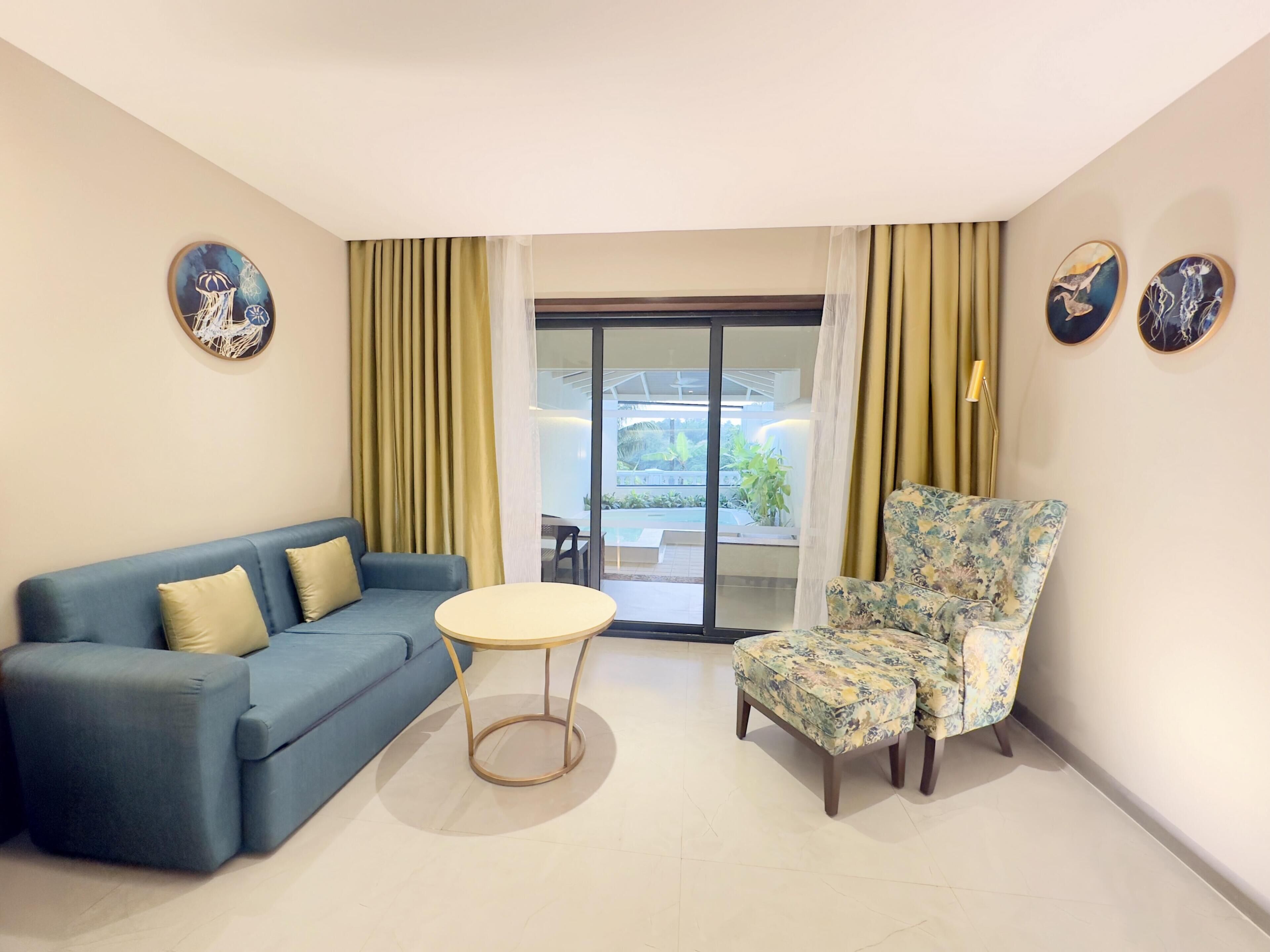 Ein Wohnzimmer mit einem blauen Sofa, einem runden Tisch, einem blumigen Sessel und Blick auf einen Außenbereich durch eine Glastür.