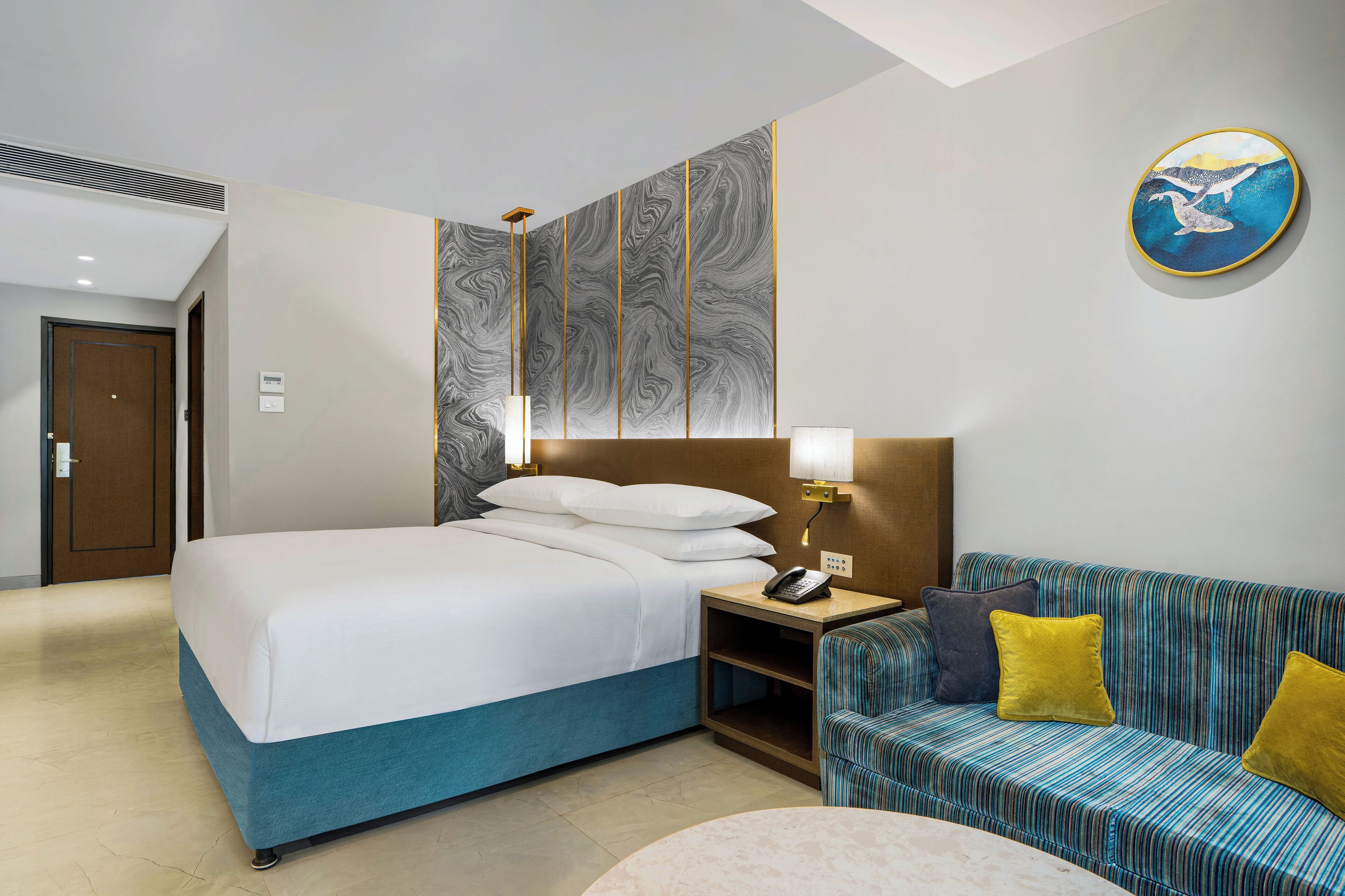 A hotel room with a bed, a sofa, a nightstand with a lamp, and a wall art piece.