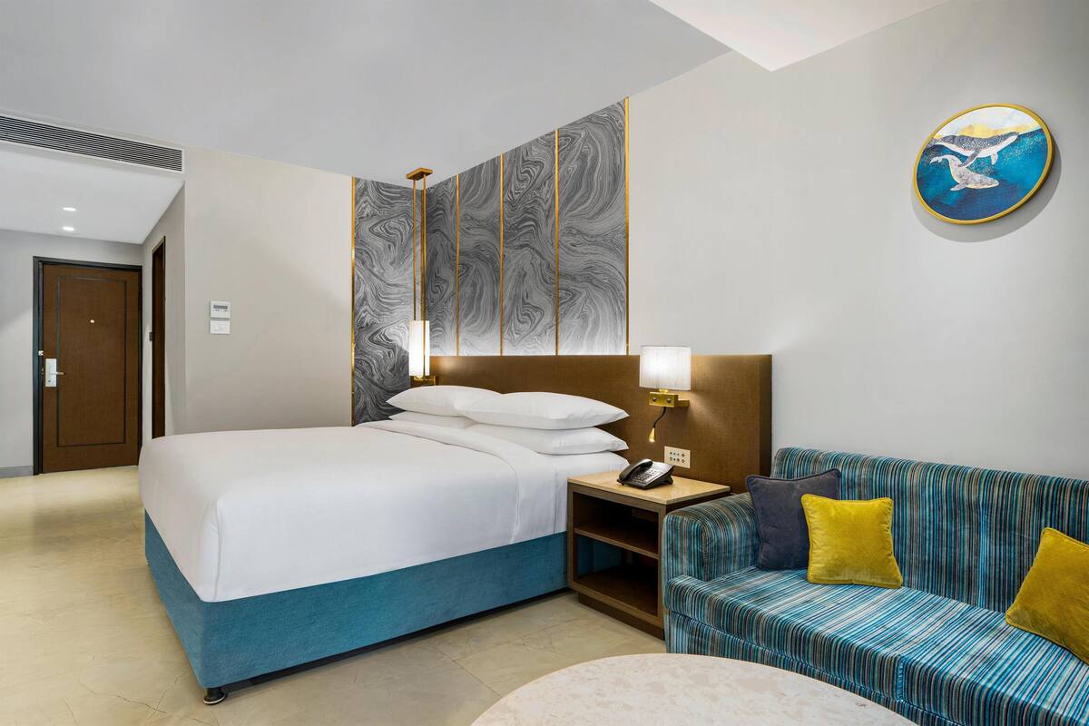 A hotel room with a bed, a sofa, a nightstand with a lamp, and a wall art piece.
