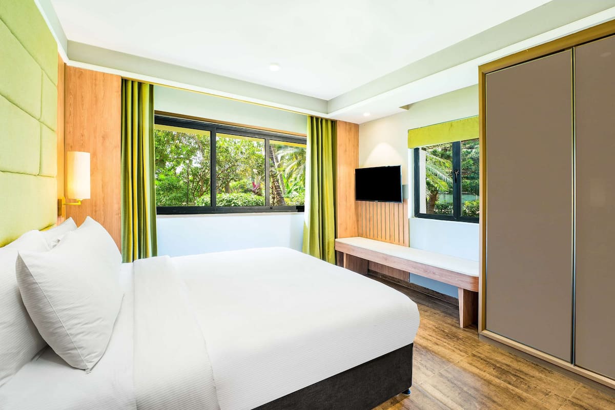 Relax in a serene space with a large bed and a view of lush greenery outside, complemented by a flat-screen TV and a cozy bench.
