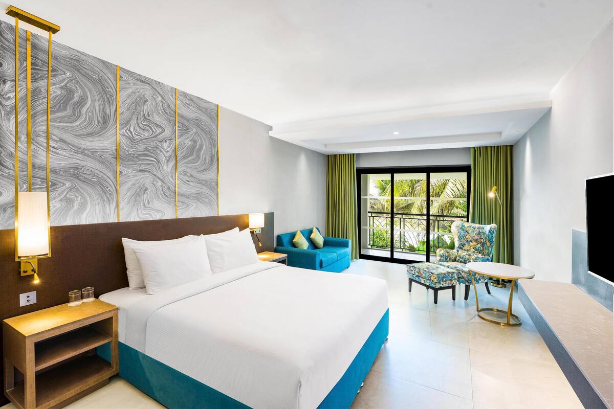 A hotel room with a large bed, a nightstand, a blue sofa, a chair, a small table, and a large window with curtains.