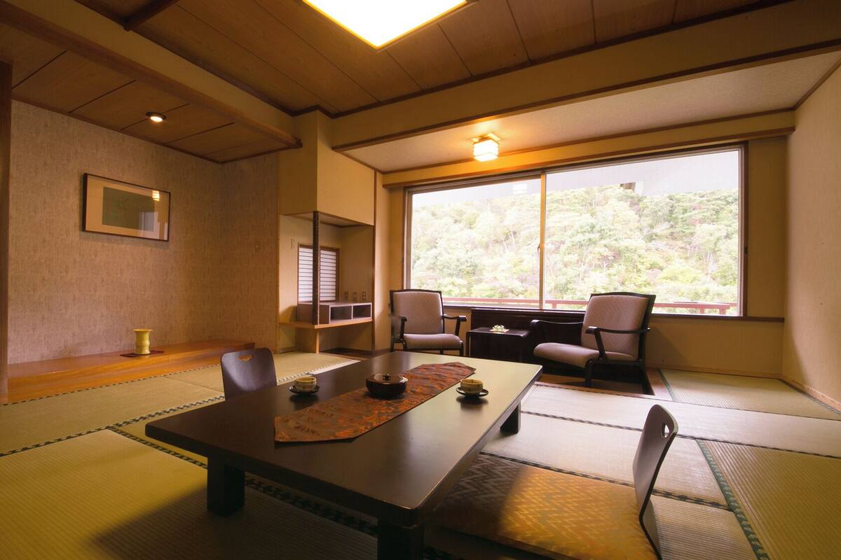 Une pièce de style japonais traditionnel, avec un sol recouvert de tatamis, une table basse, des chaises, une fenêtre donnant sur un paysage verdoyant et un luminaire au plafond.