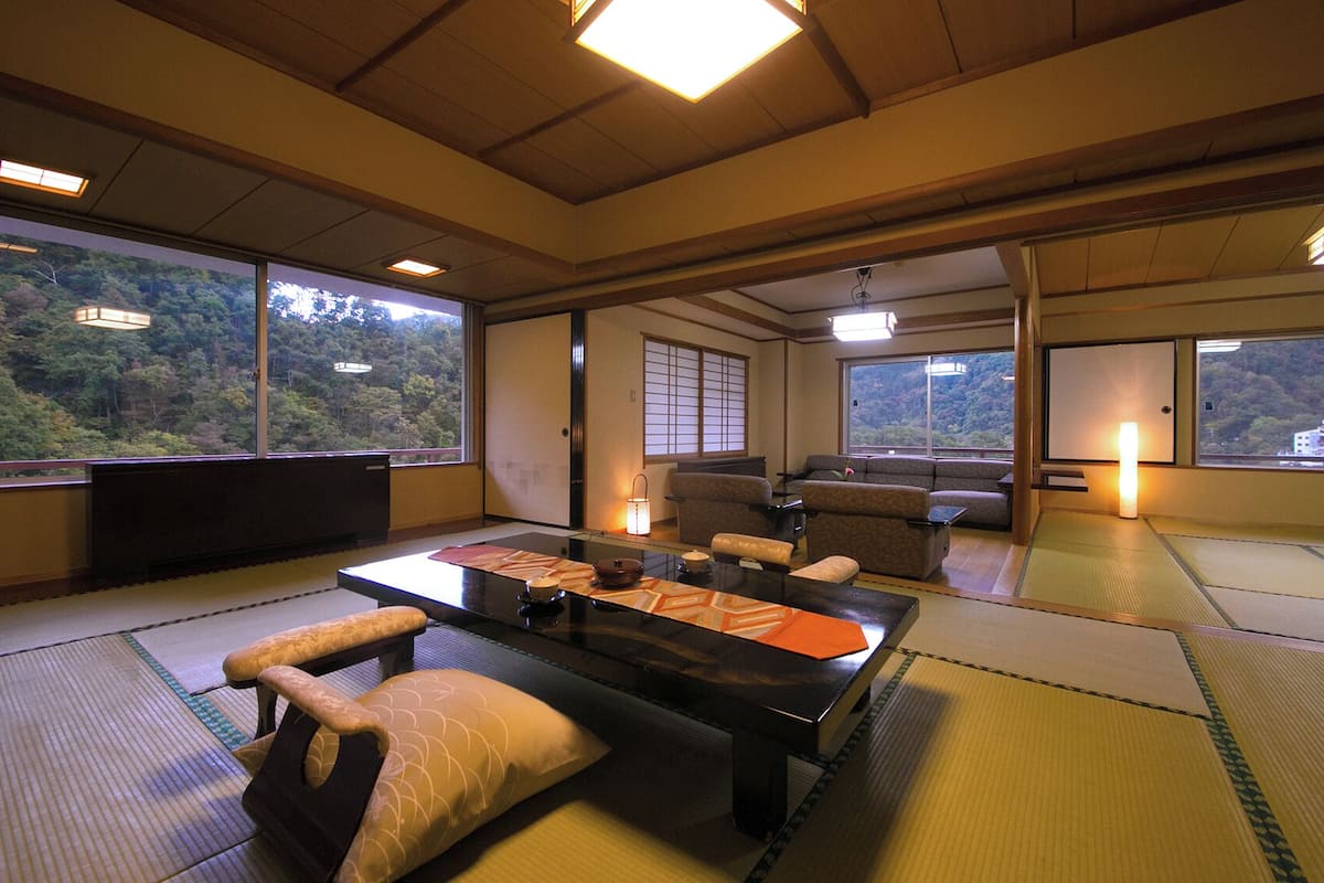 Um quarto tradicional com esteiras de tatame, uma mesa baixa, almofadas e vista para uma encosta arborizada.