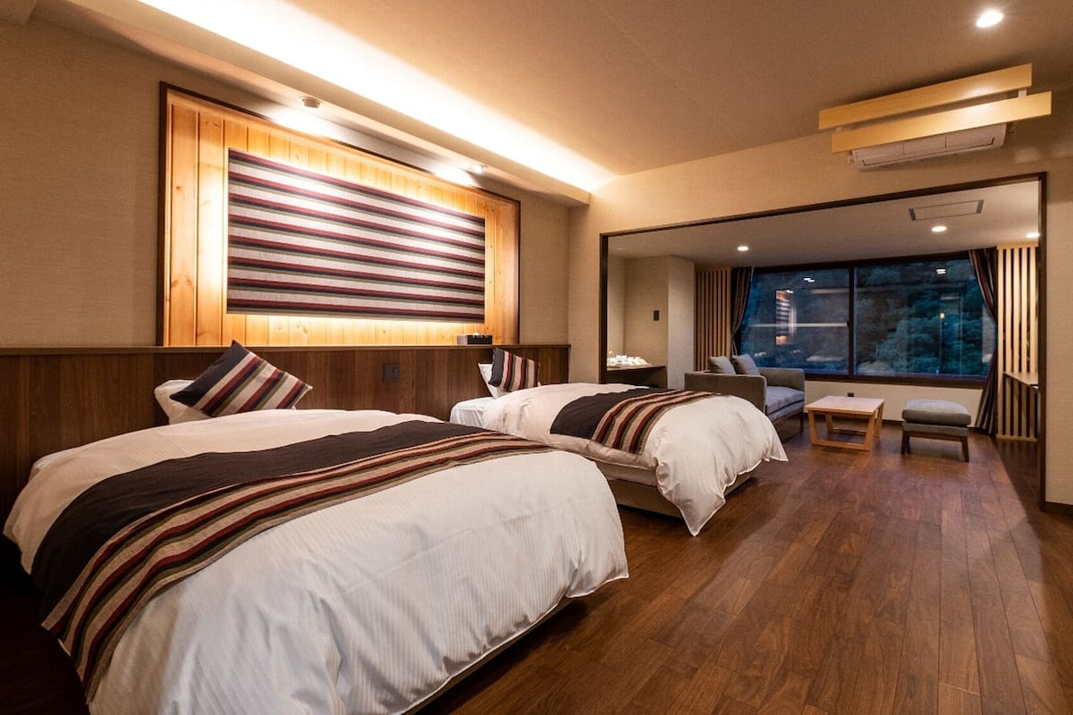 Une chambre d’hôtel avec deux lits, une tête de lit en bois, une grande fenêtre donnant sur un beau paysage et un petit coin salon.