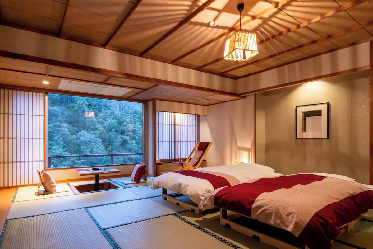 Une pièce de style japonais traditionnel, avec un sol recouvert de tatamis, un futon servant de lit, un plafond en bois et une vue sur une forêt, visible à travers de grandes fenêtres.