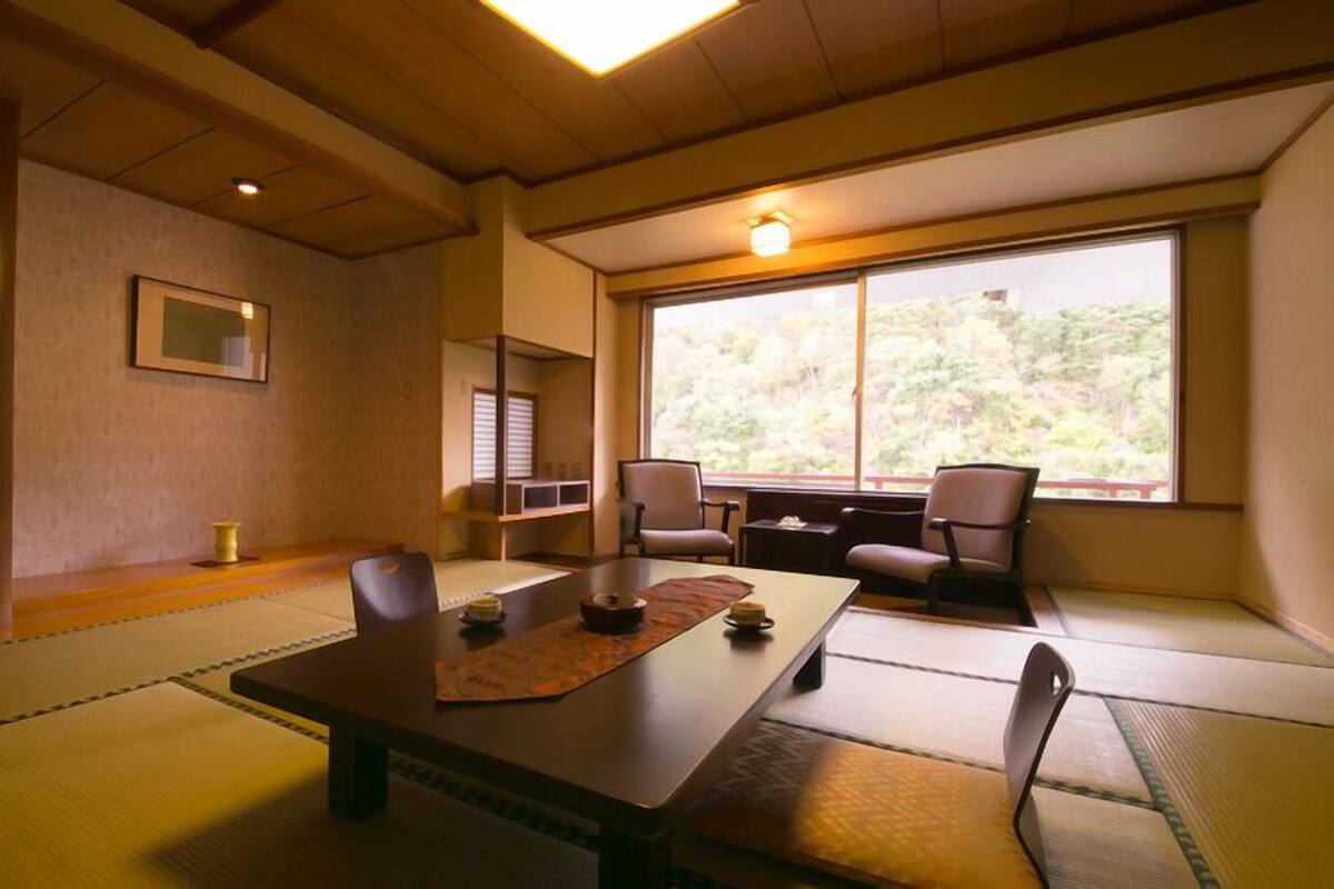 Une pièce de style japonais traditionnel, avec un sol recouvert de tatamis, une table basse, des chaises, une fenêtre donnant sur un paysage verdoyant et un tableau encadré accroché au mur.