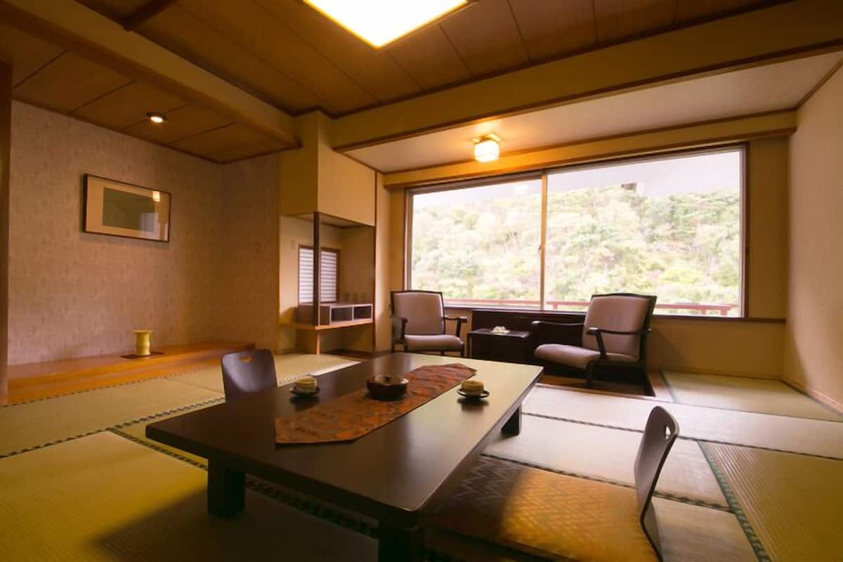 Um quarto japonês tradicional com tatames, uma mesa baixa, cadeiras, uma janela com vista para o verde e um quadro na parede.