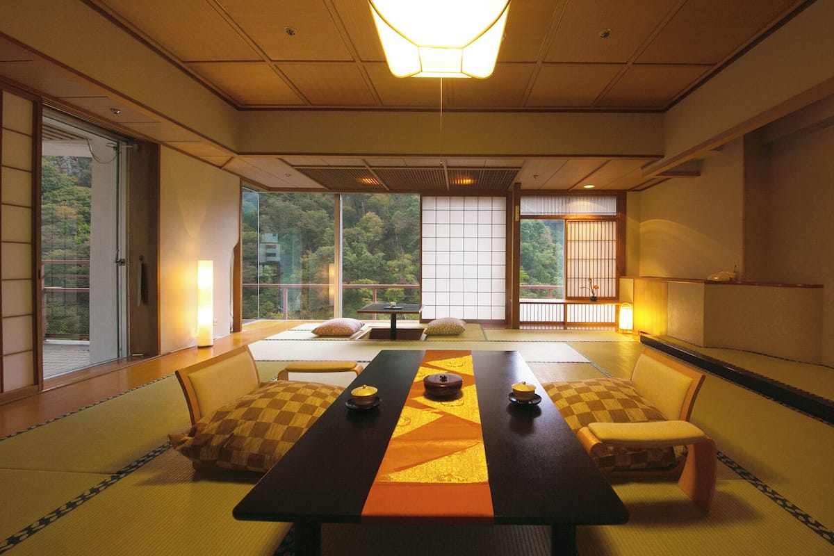 Une pièce de style japonais traditionnel, avec un sol recouvert de tatamis, une table basse avec des coussins, des portes coulissantes et une vue sur une zone boisée.