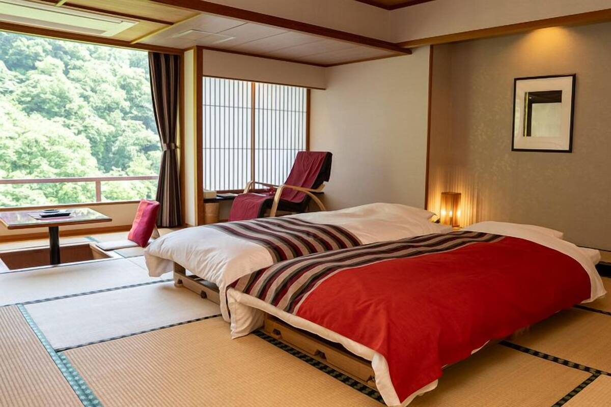 Une chambre d’hôtel avec un sol recouvert de tatamis, un lit avec une couverture à rayures rouges et blanches, un bureau avec une chaise, une fenêtre avec des stores et un tableau encadré au mur.
