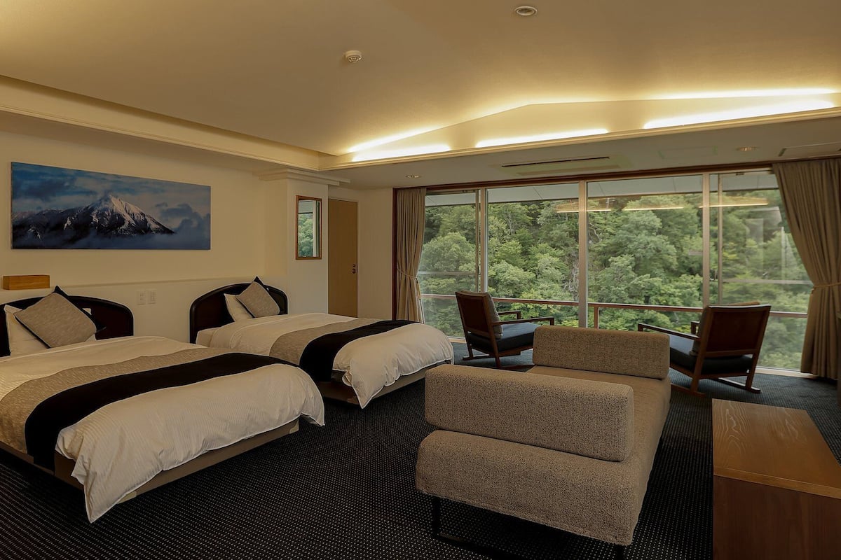 Une chambre d’hôtel avec deux lits, un canapé, un bureau et une grande fenêtre donnant sur la forêt.