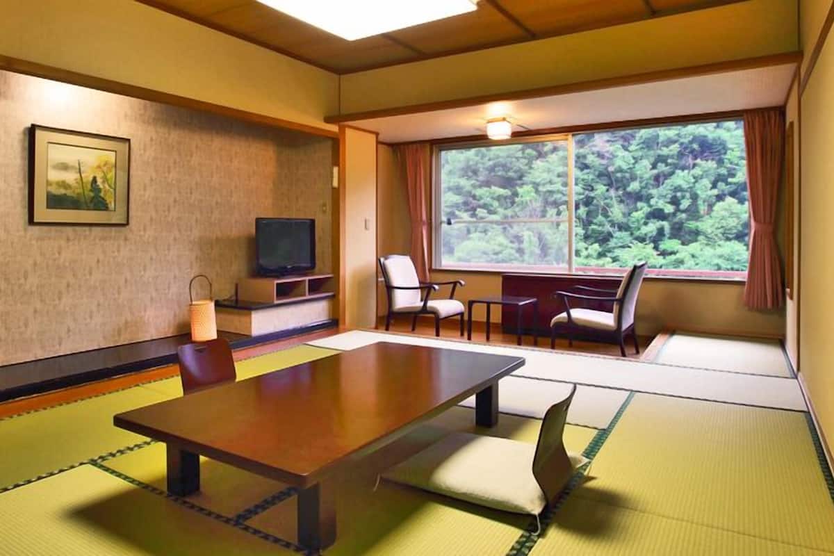 Une pièce de style japonais traditionnel, avec un sol recouvert de tatamis, une table basse, des sièges, un téléviseur et une fenêtre donnant sur un paysage verdoyant.