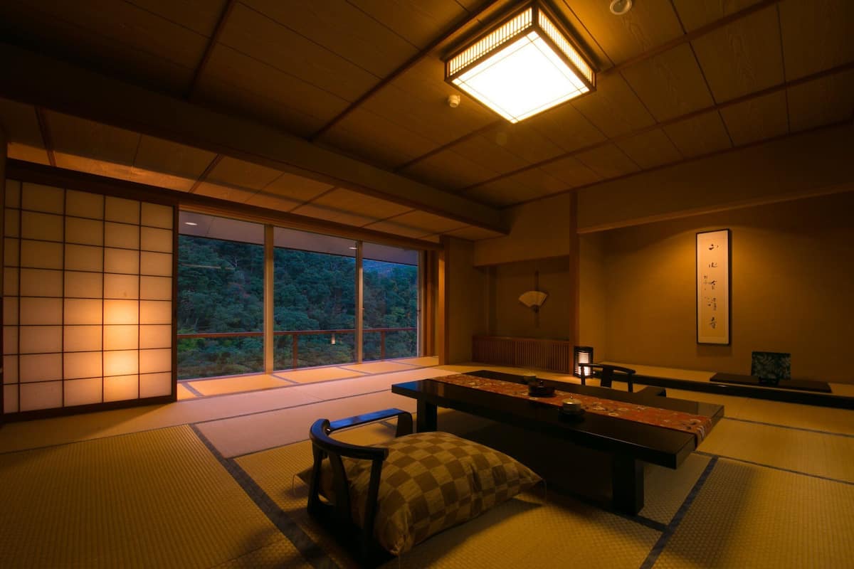 Une pièce de style japonais traditionnel, avec un sol recouvert de tatamis, une table basse et une vue sur une forêt, visible à travers de grandes fenêtres.
