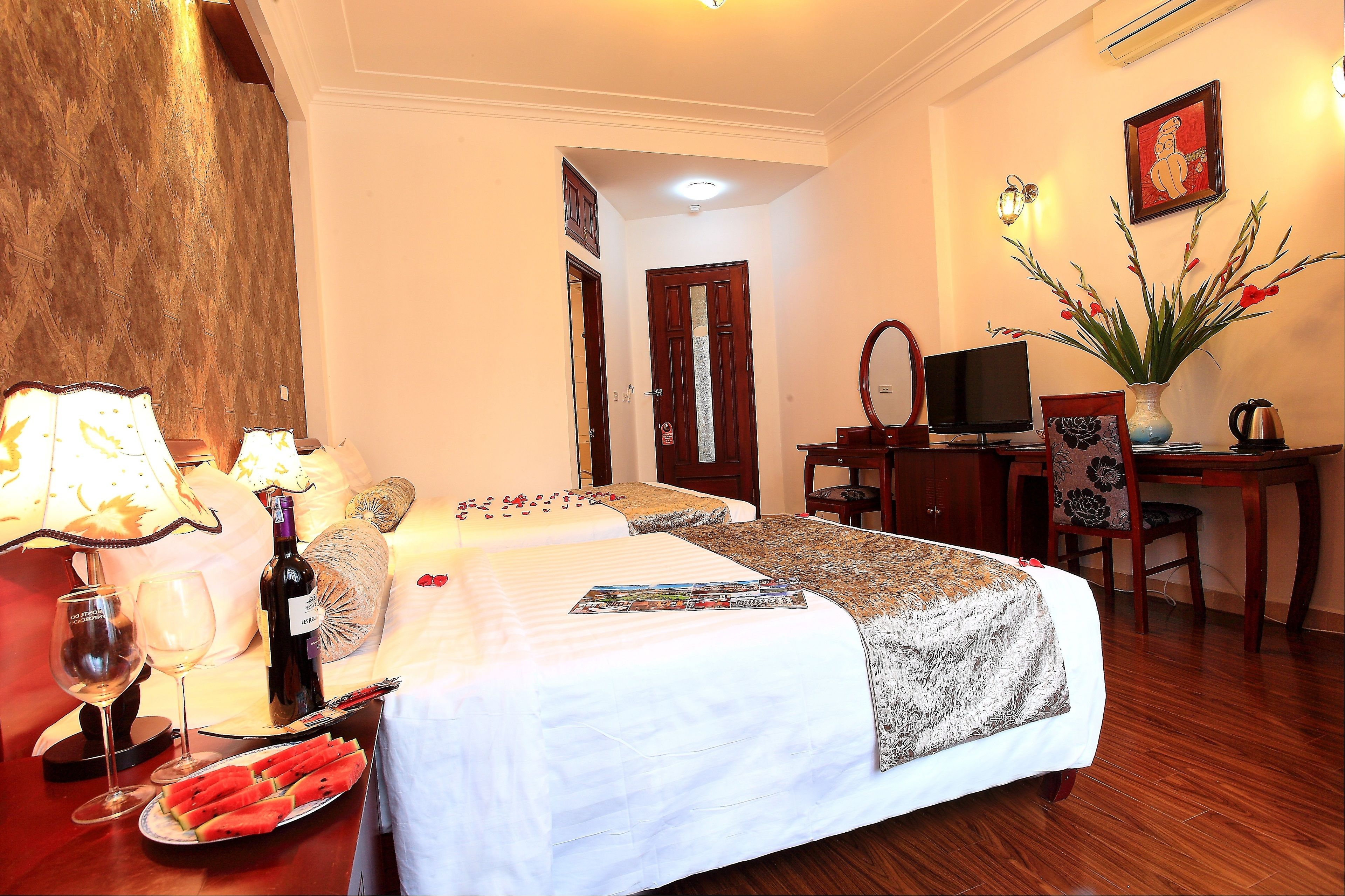 A hotel room with a bed, a desk, a chair, a television, a vase with flowers, and a wine bottle.