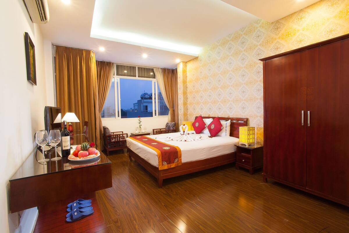A hotel room with a bed, a wooden wardrobe, a desk with a lamp, and a window with curtains.