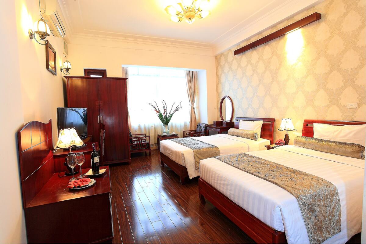 A hotel room with two beds, wooden flooring, a desk with a lamp, a mirror, and a window with curtains.