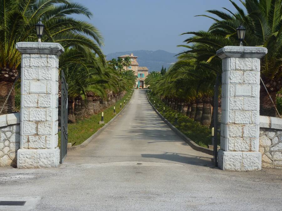 A paved road flanked by stone pillars with inscriptions, leading to a building with a tiled roof and a view of mountains in the distance.