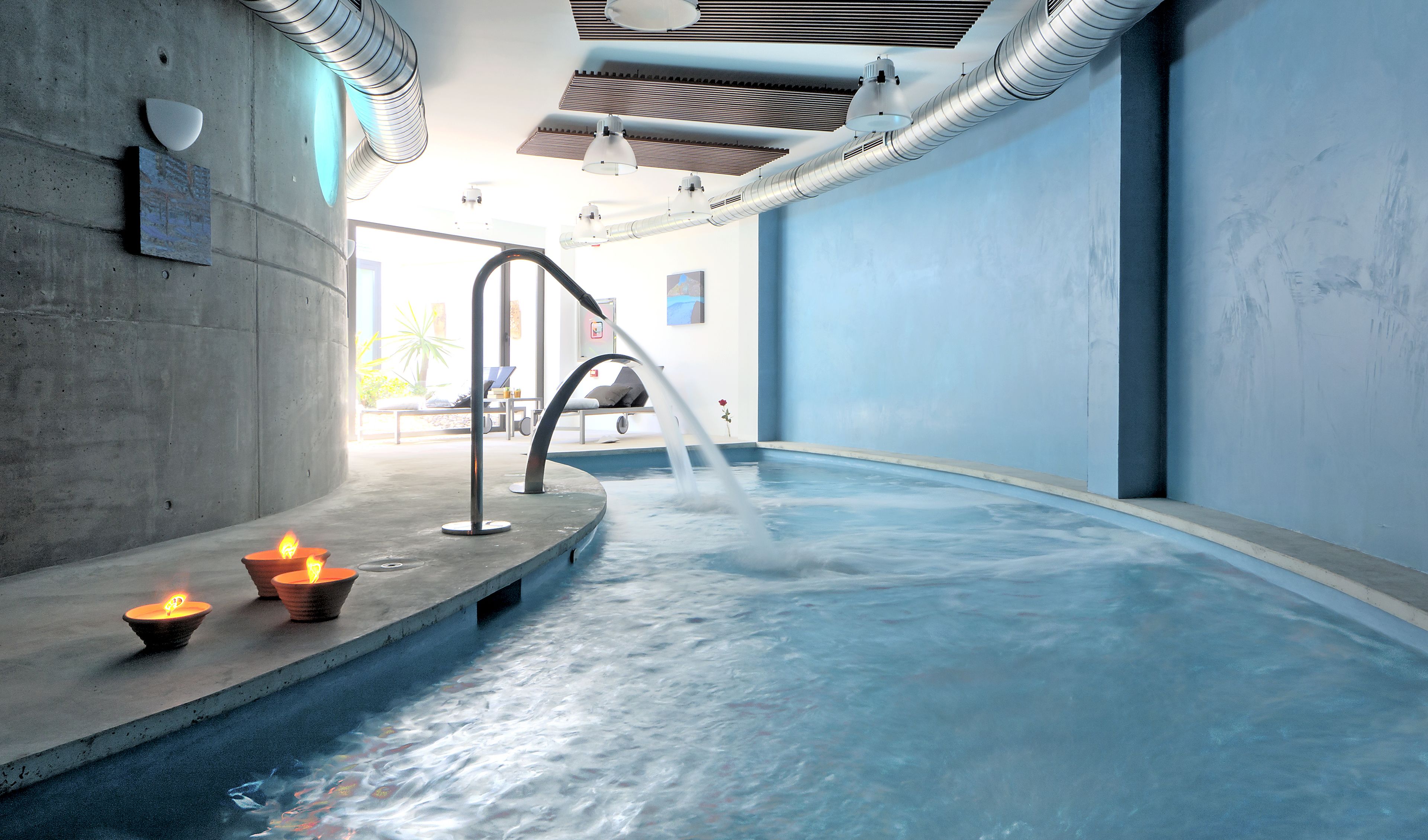 Une piscine intérieure moderne agrémentée d’un élément aquatique décoratif, un mur en béton orné d’une fresque et un plafond doté de conduits de ventilation.