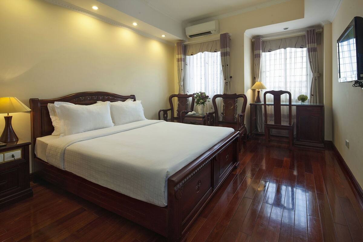 A neatly arranged bedroom with a wooden bed, bedside lamps, a desk with a chair, and a window with curtains.