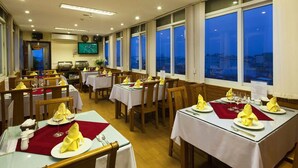 A dining area with tables set for service, a television mounted on the wall, and large windows with a view of the city at dusk. - Lucky 3 Hotel & Travel  (Hanoi)