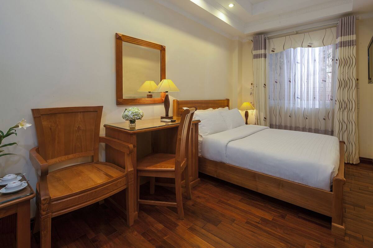 A hotel room with a wooden bed, a wooden desk with two lamps, a chair, and a small table with a vase of flowers.