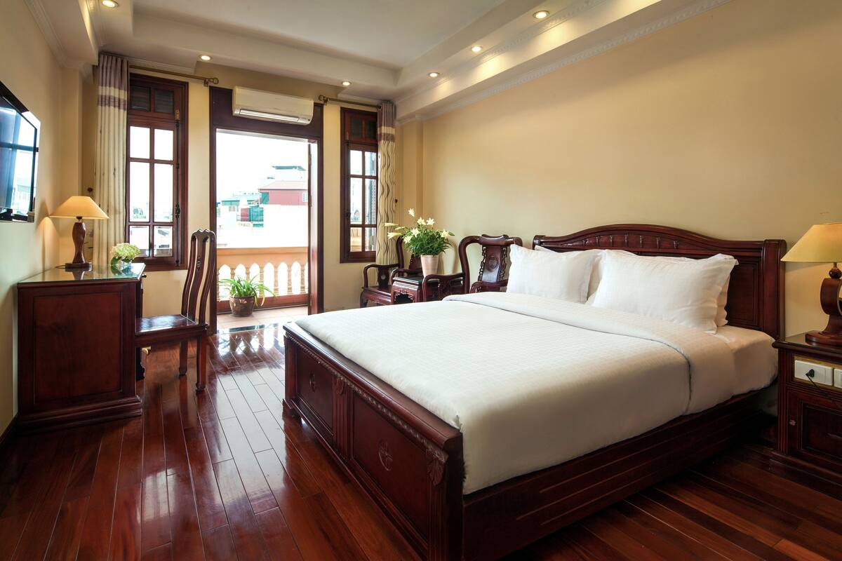 A spacious bedroom with a large bed, wooden flooring, a potted plant, and a view of the outside through a window.
