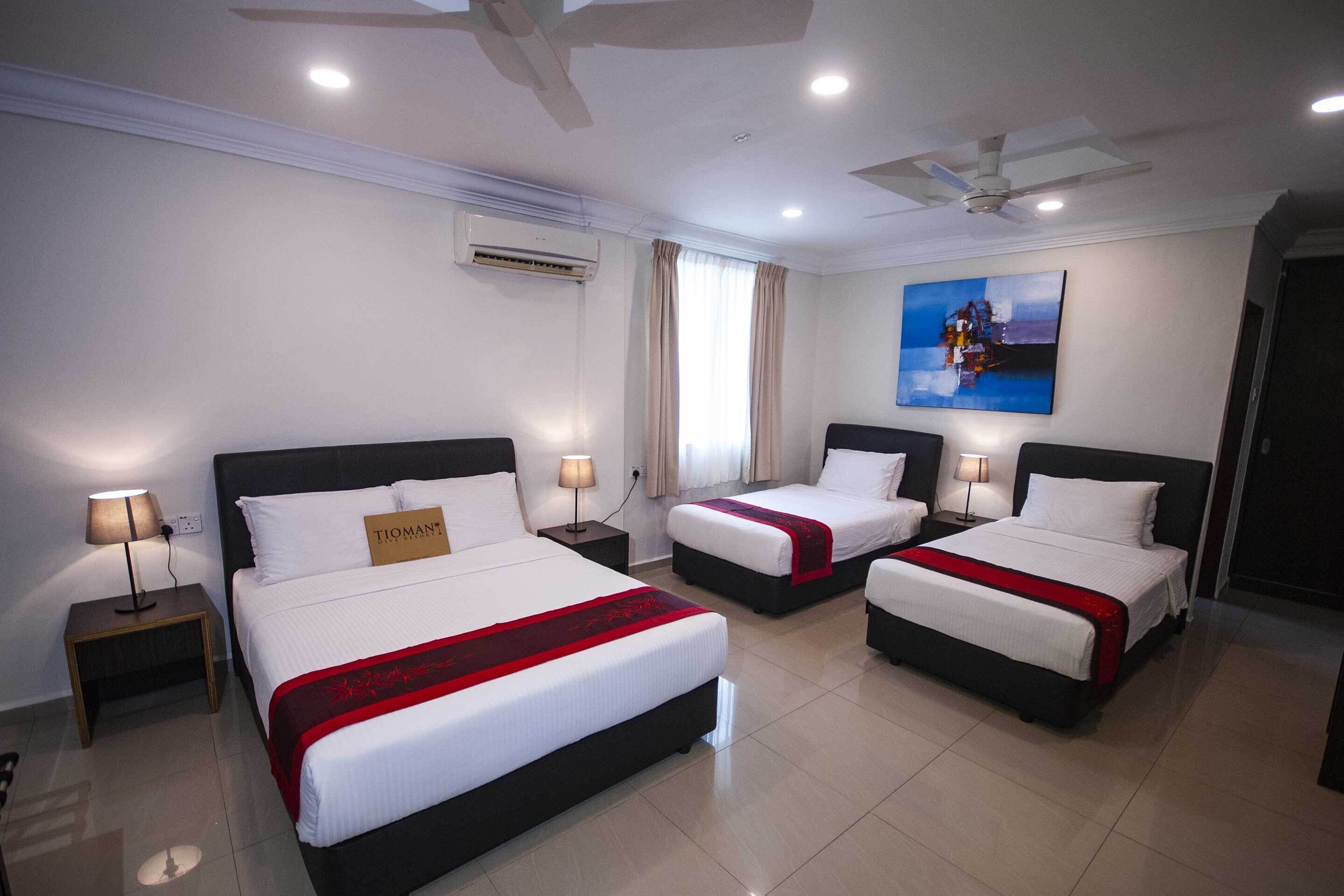 A hotel room with three beds, each with a red and white bedspread, white pillows, and a small bedside table with a lamp.