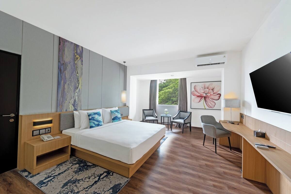 Ein modernes Hotelzimmer mit einem großen Bett, Nachttischen, einem Schreibtisch, einem Sessel, einem Fernseher und einem Fenster mit Blick auf Grünflächen.