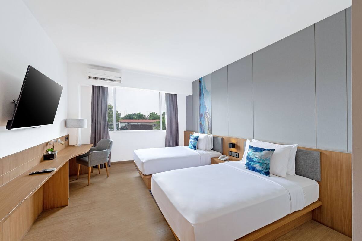 Ein Hotelzimmer mit zwei Betten, einem Schreibtisch, einem Fernseher und einem Fenster mit Vorhängen.
