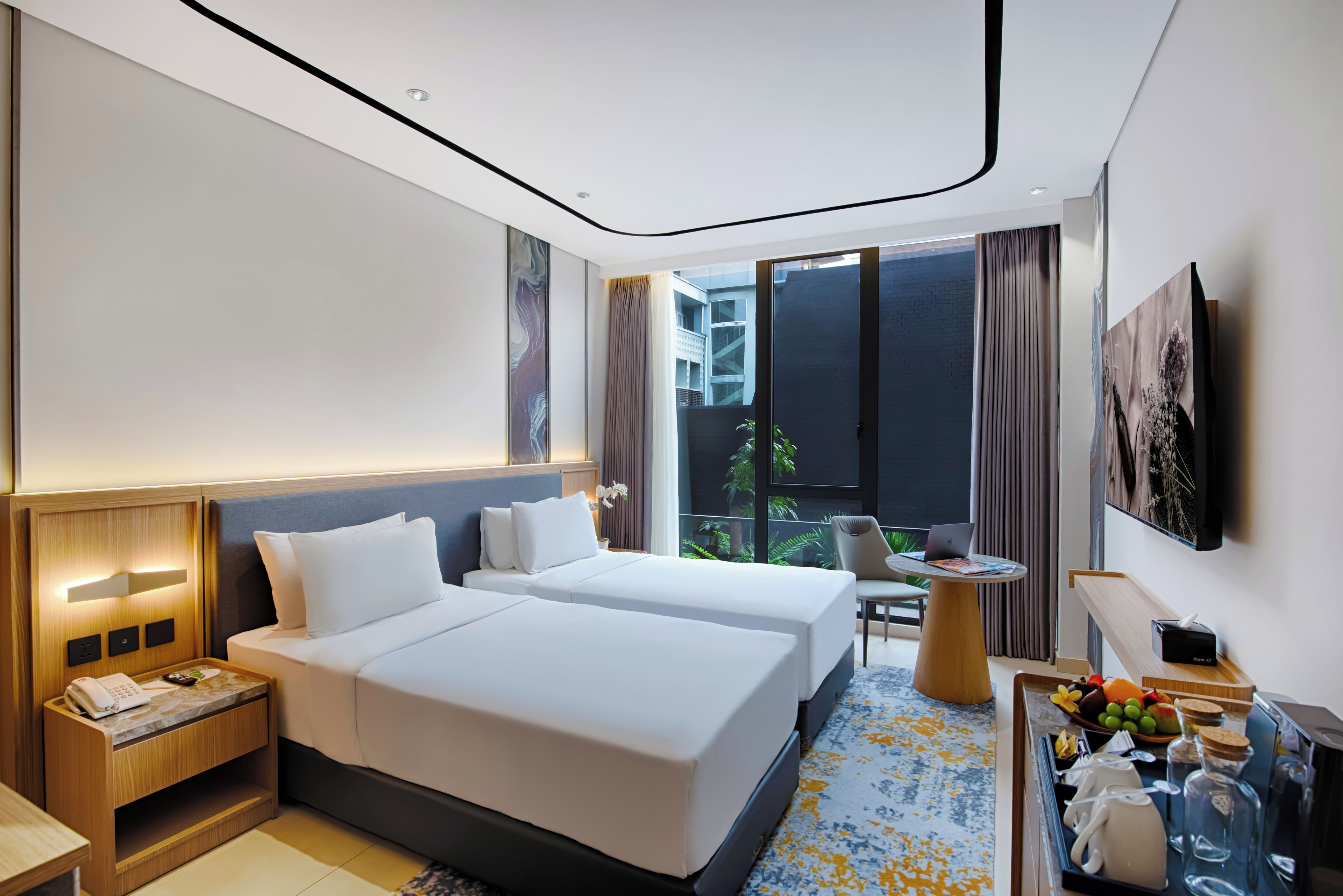 A hotel room with two beds, a desk, a chair, a small table, a TV, and a window with a view of buildings.