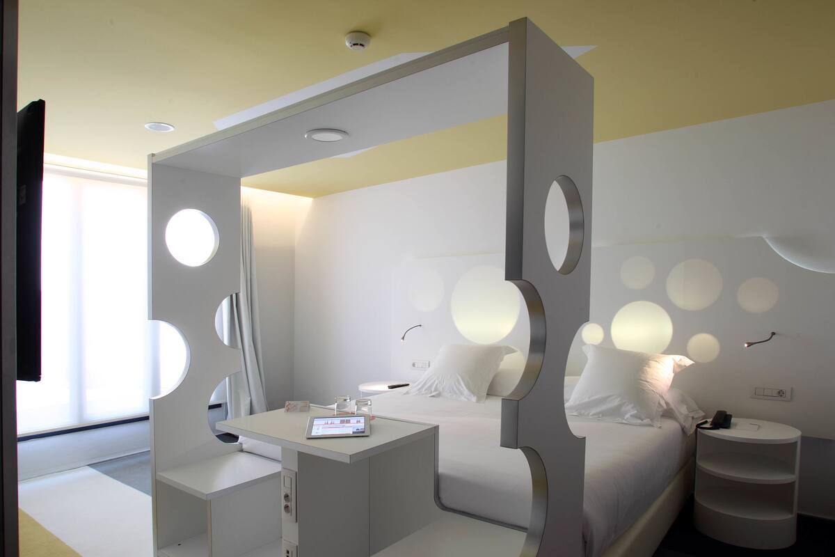 Una habitación de hotel moderna con cama, mesita de noche y un pequeño escritorio con una computadora.