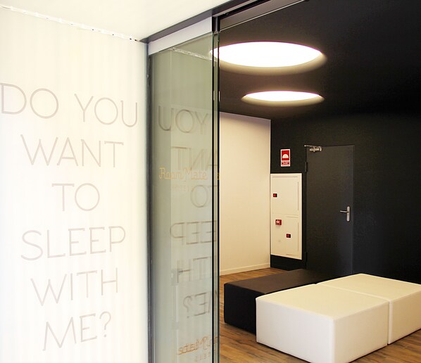 Una habitación de hotel moderna con un separador de vidrio, una cama blanca y un pequeño rincón de descanso.