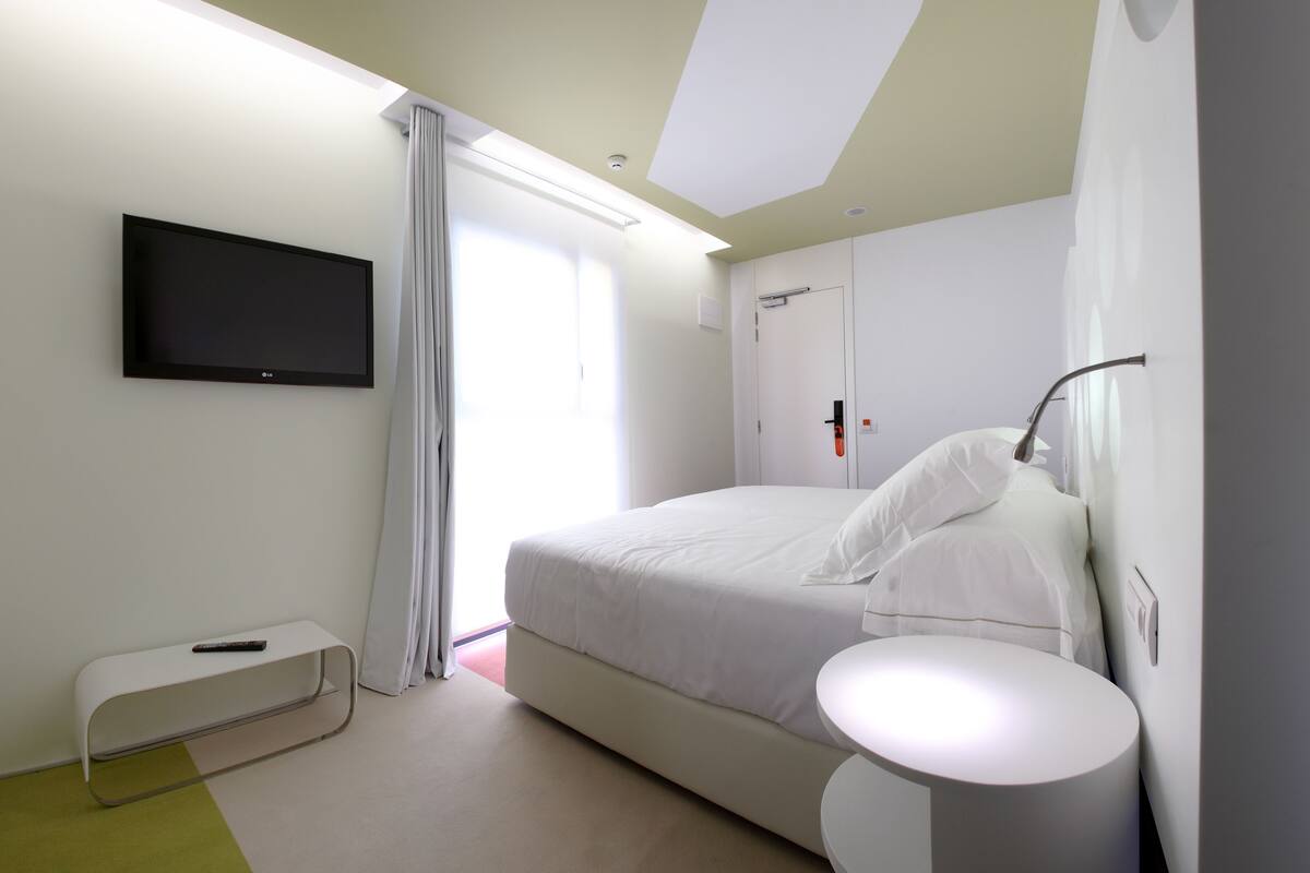 Una habitación de hotel moderna con una cama, un televisor montado en la pared, una mesita de noche y una pequeña mesa redonda.