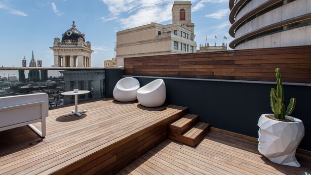 Terraço no telhado com piso de madeira, duas poltronas brancas, uma mesa pequena e um cacto em vaso.