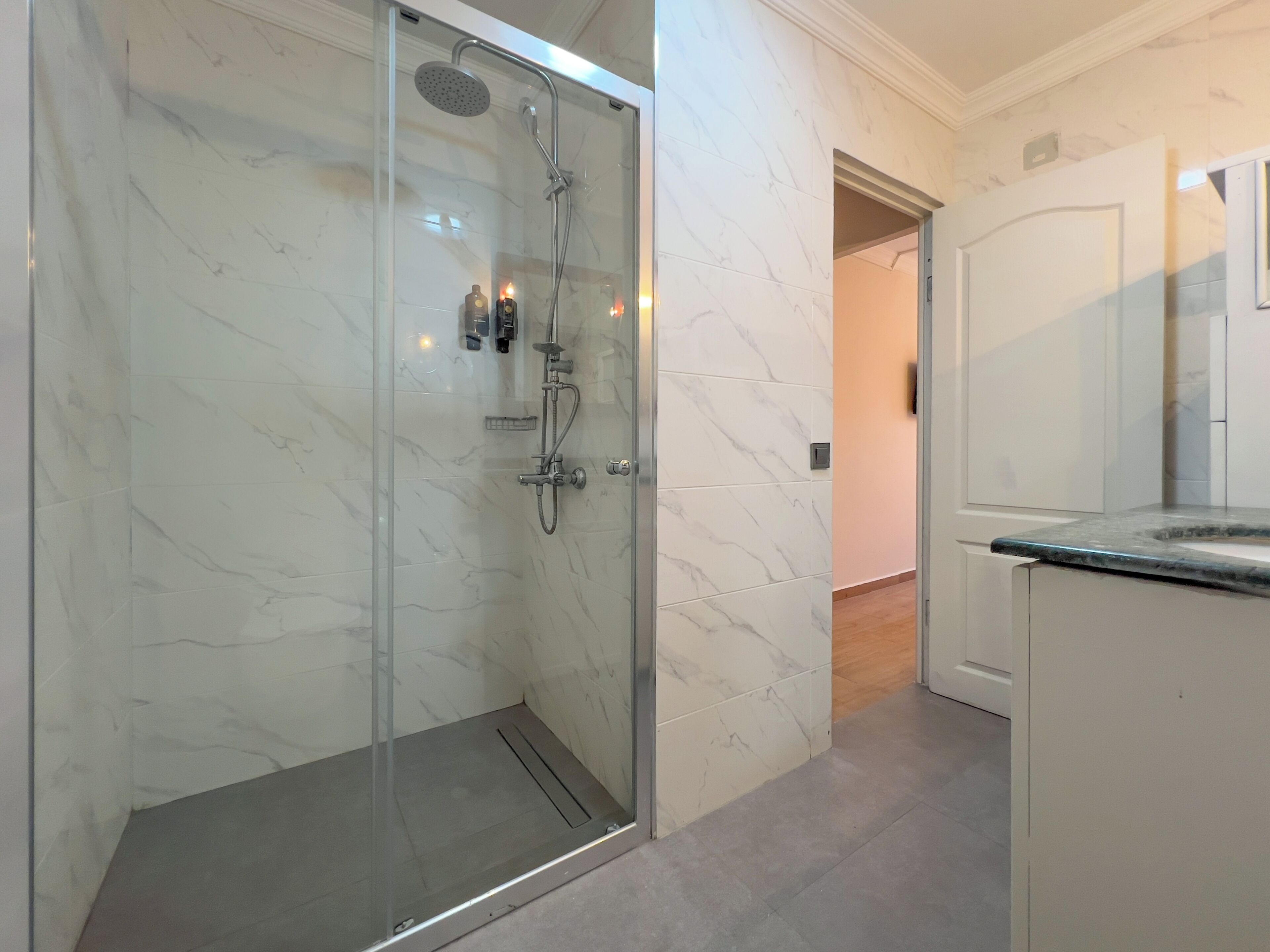 A modern bathroom with a glass-enclosed shower, marble walls, and a wooden floor.