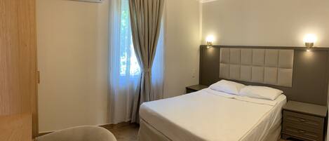Una habitación de hotel con cama, mesitas de noche, aire acondicionado, una silla y ventana con cortinas.