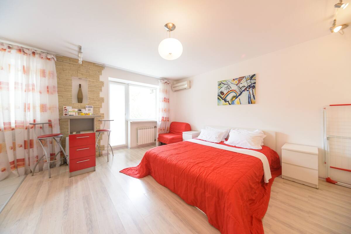 Un dormitorio con cama, escritorio y silla. Hay una ventana con cortinas, un equipo de aire acondicionado y un cuadro en la pared.