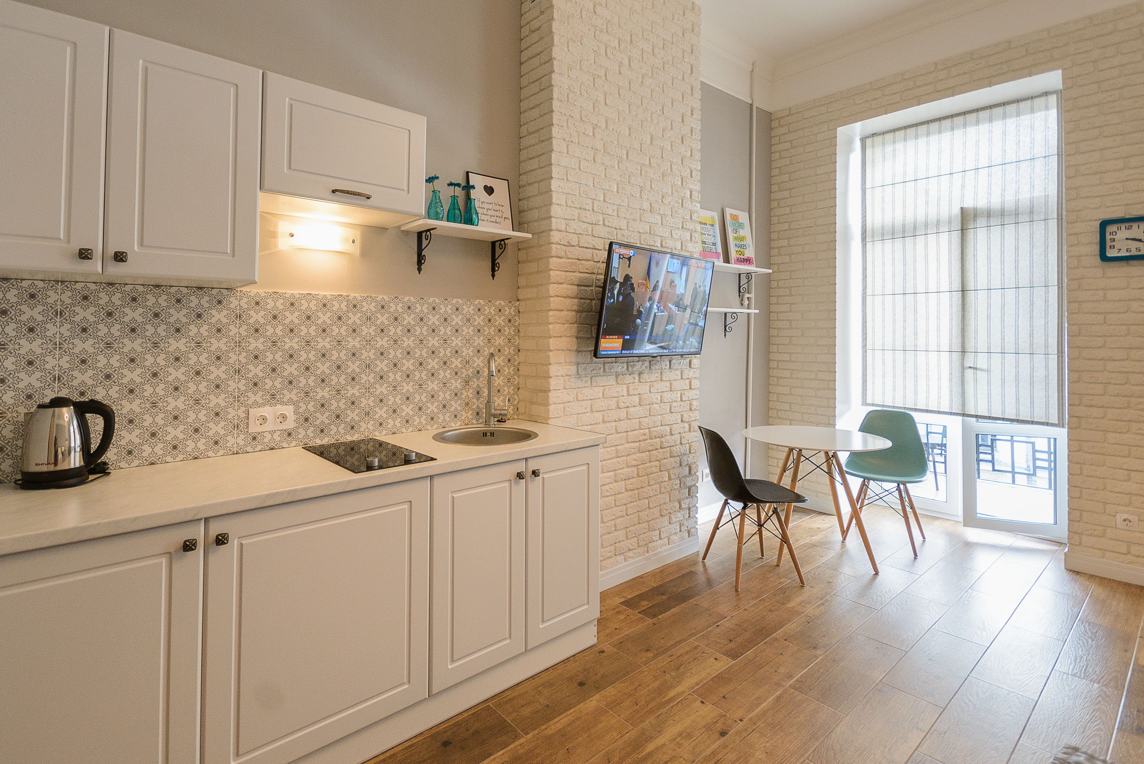 A kitchen with white cabinets, a sink, and a kettle on the counter. There is a television mounted on the wall, and a dining area with a table and chairs.