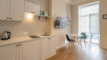 A kitchen with white cabinets, a sink, and a kettle on the counter. There is a television mounted on the wall, and a dining area with a table and chairs.
