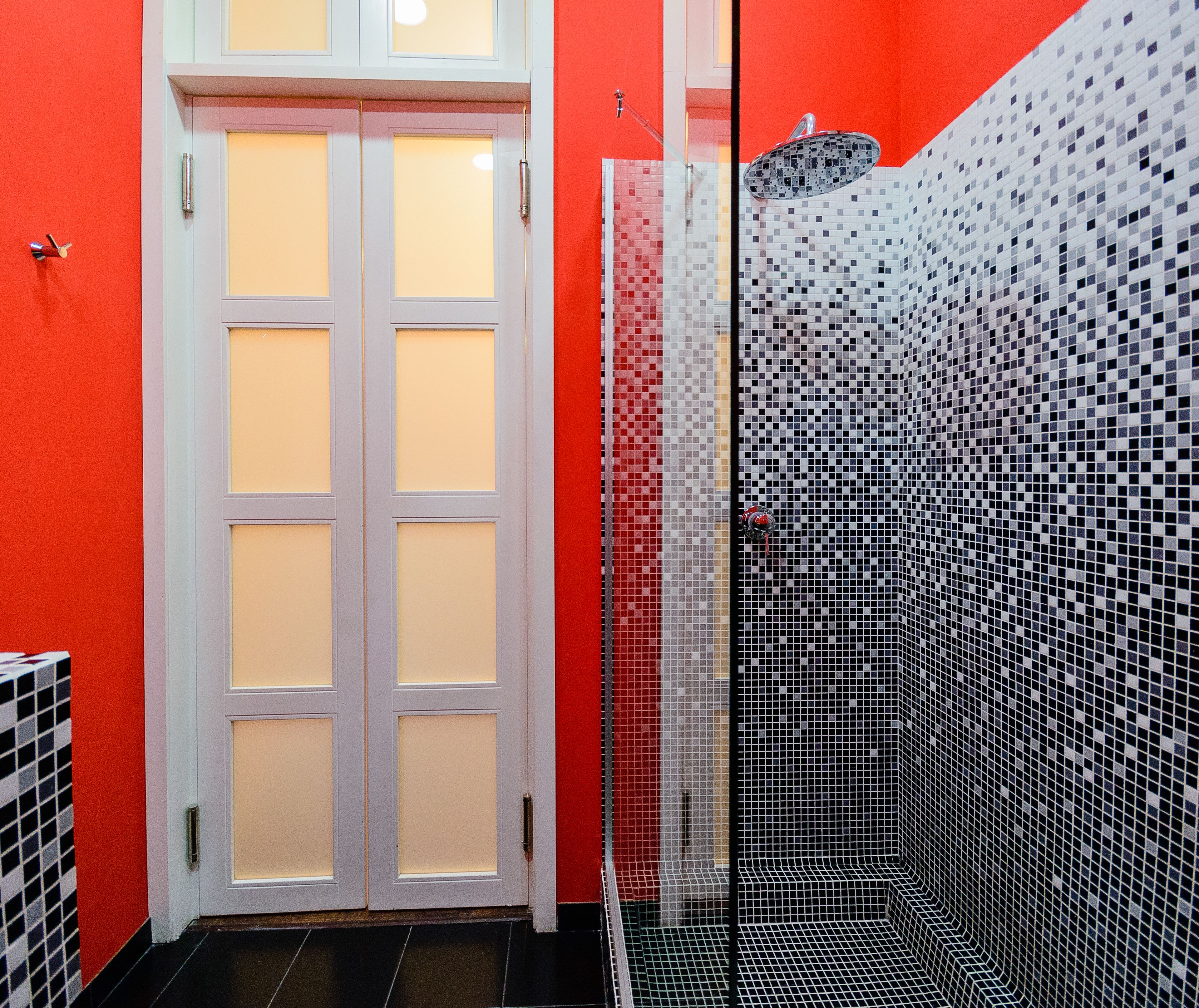Um banheiro com parede vermelha, porta branca e área de chuveiro com azulejos de mosaico preto e branco.