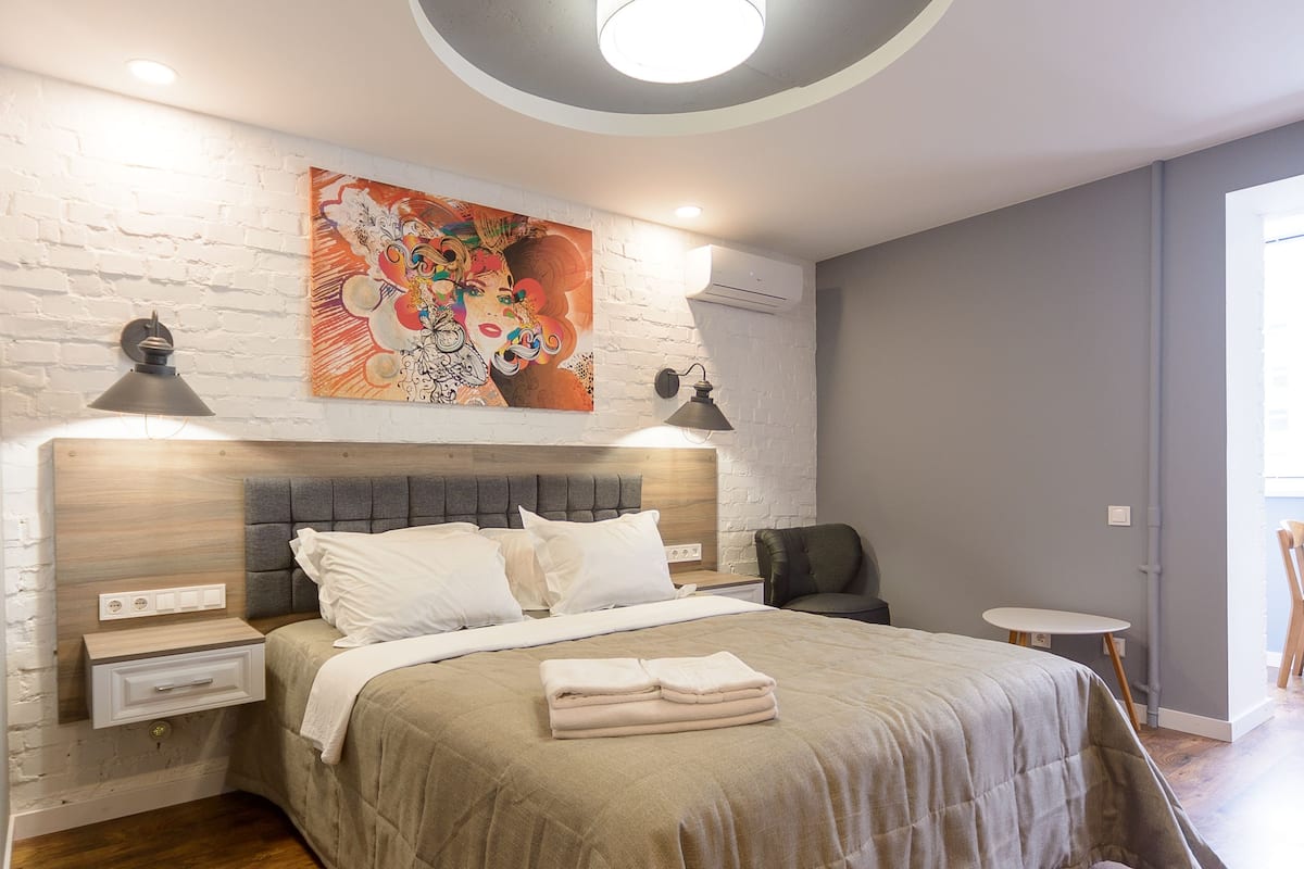 Un dormitorio moderno con cabecera de madera, una cama grande con ropa de cama color beige, una lámpara de techo redonda y una mesita pequeña.