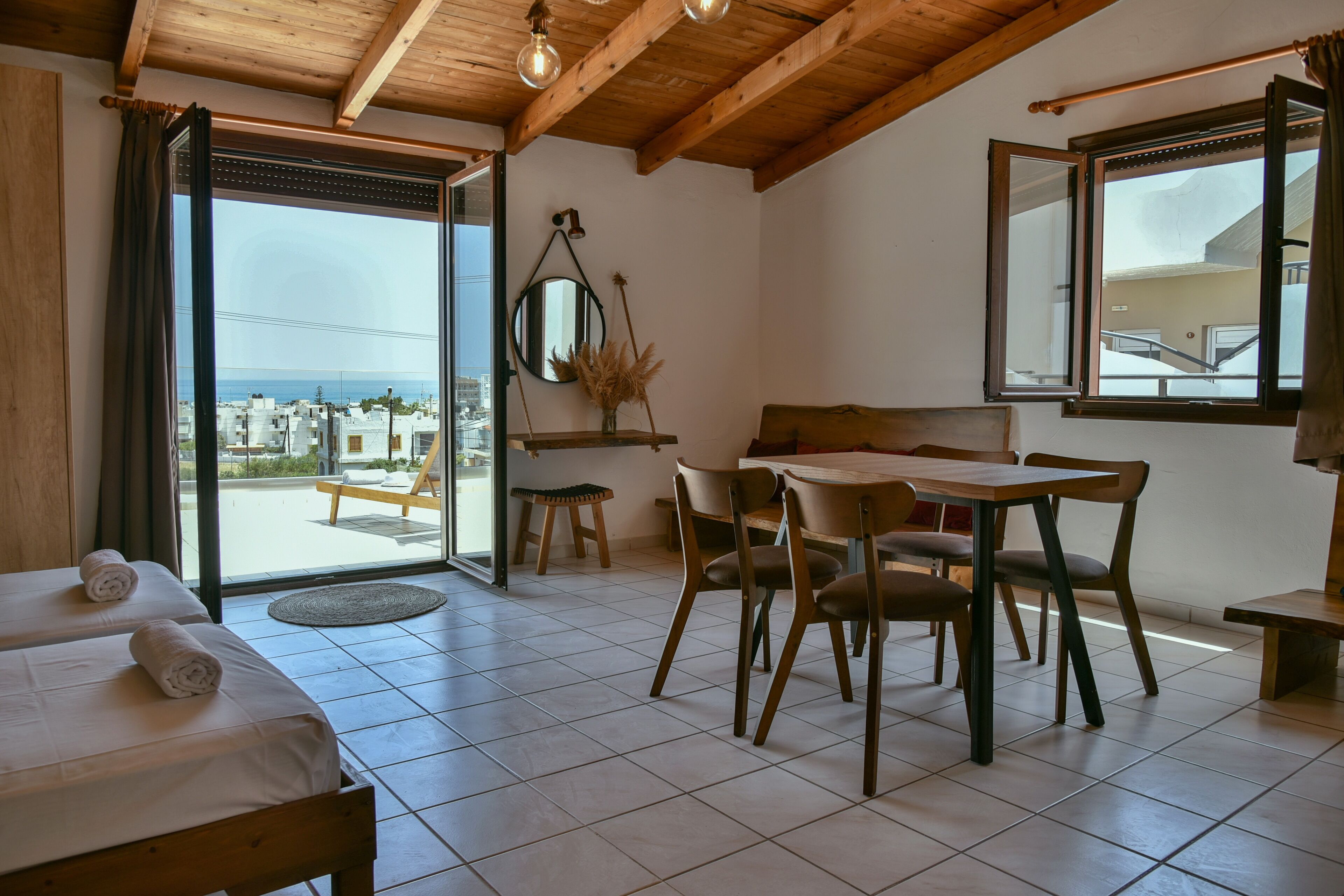 Une chambre comprenant un lit, une table à manger avec des chaises, un petit tabouret, un miroir et une vue sur la mer par une grande fenêtre.