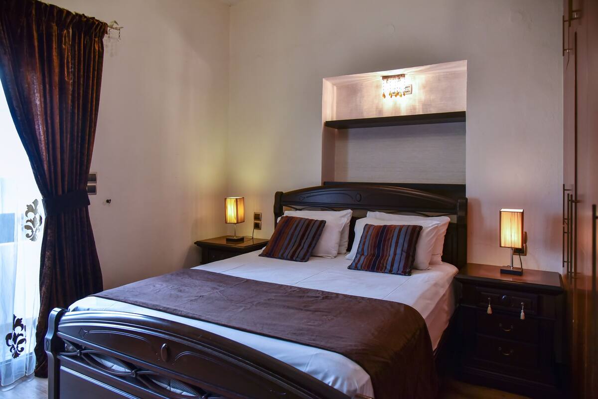 A hotel room with a large bed, two bedside tables with lamps, a headboard, and a window with curtains.