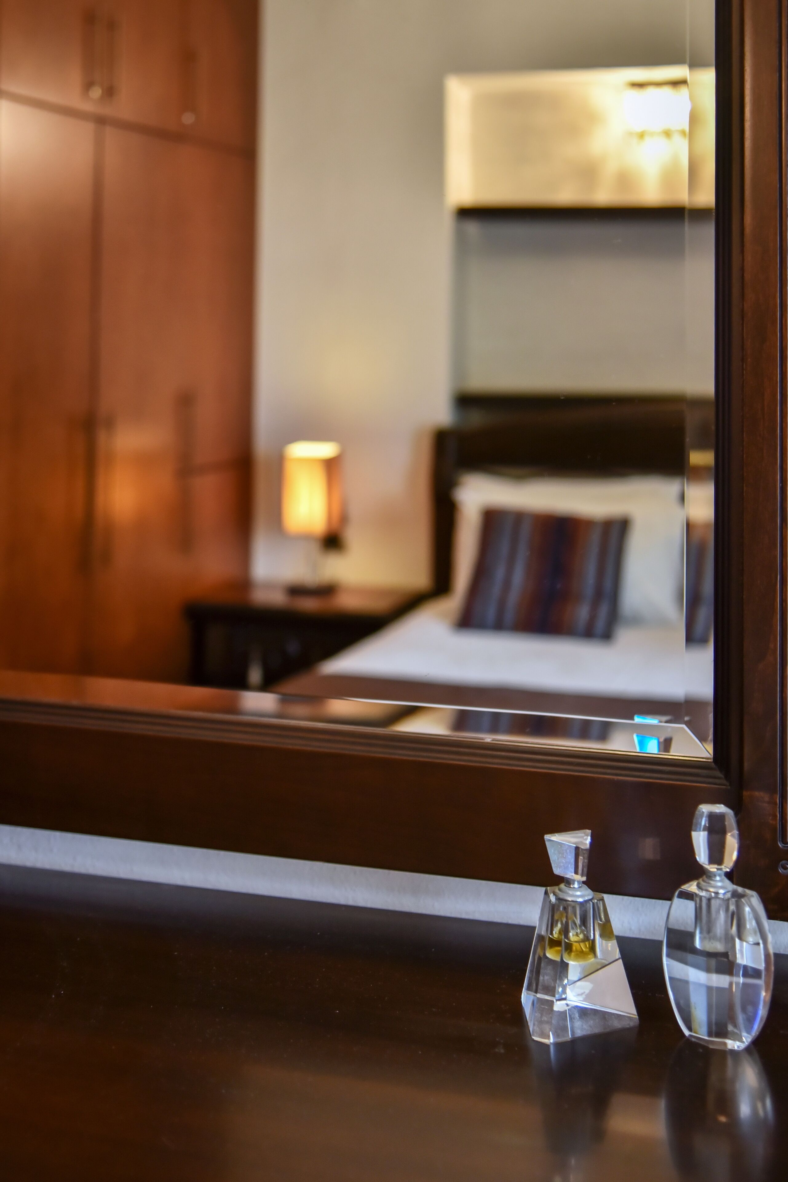 A hotel room with a wooden wardrobe, a bedside lamp, a bed with a striped pillow, and two glass perfume bottles on the nightstand.