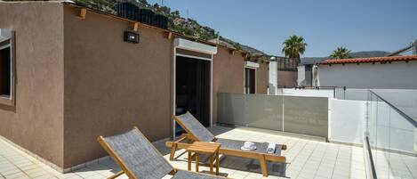 A rooftop terrace with two wooden lounge chairs, a small building, and a view of the surrounding landscape.