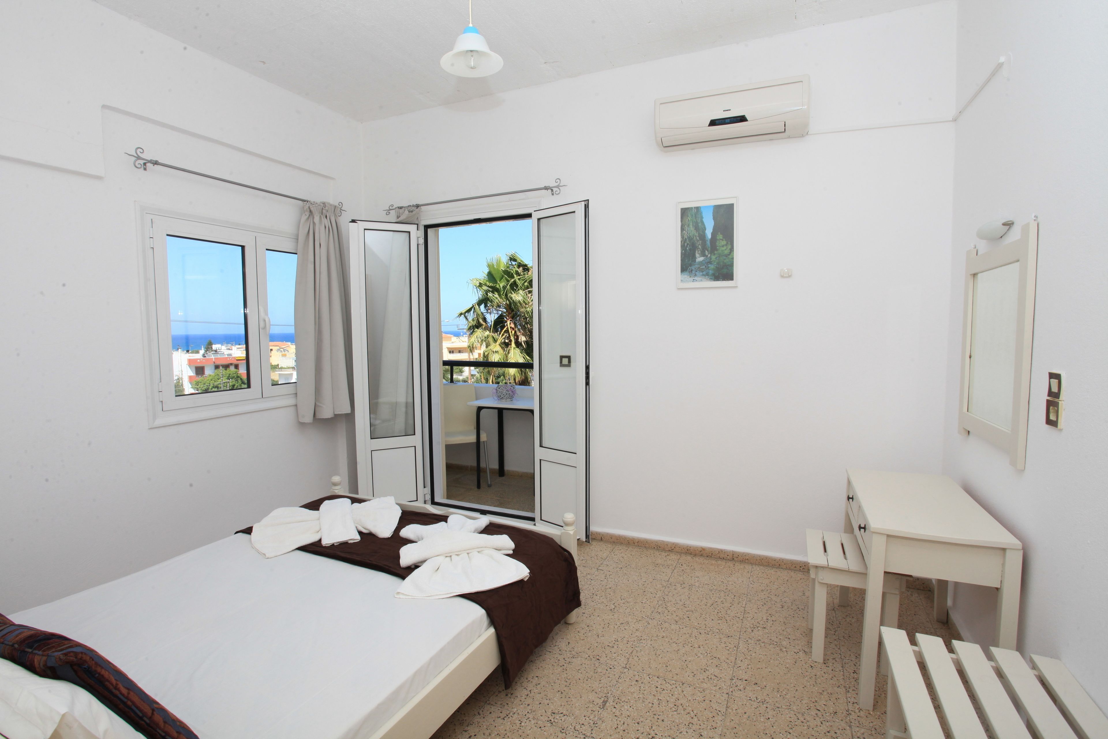 A hotel room with a bed, a small desk, a mirror, and a view of the sea through an open door.