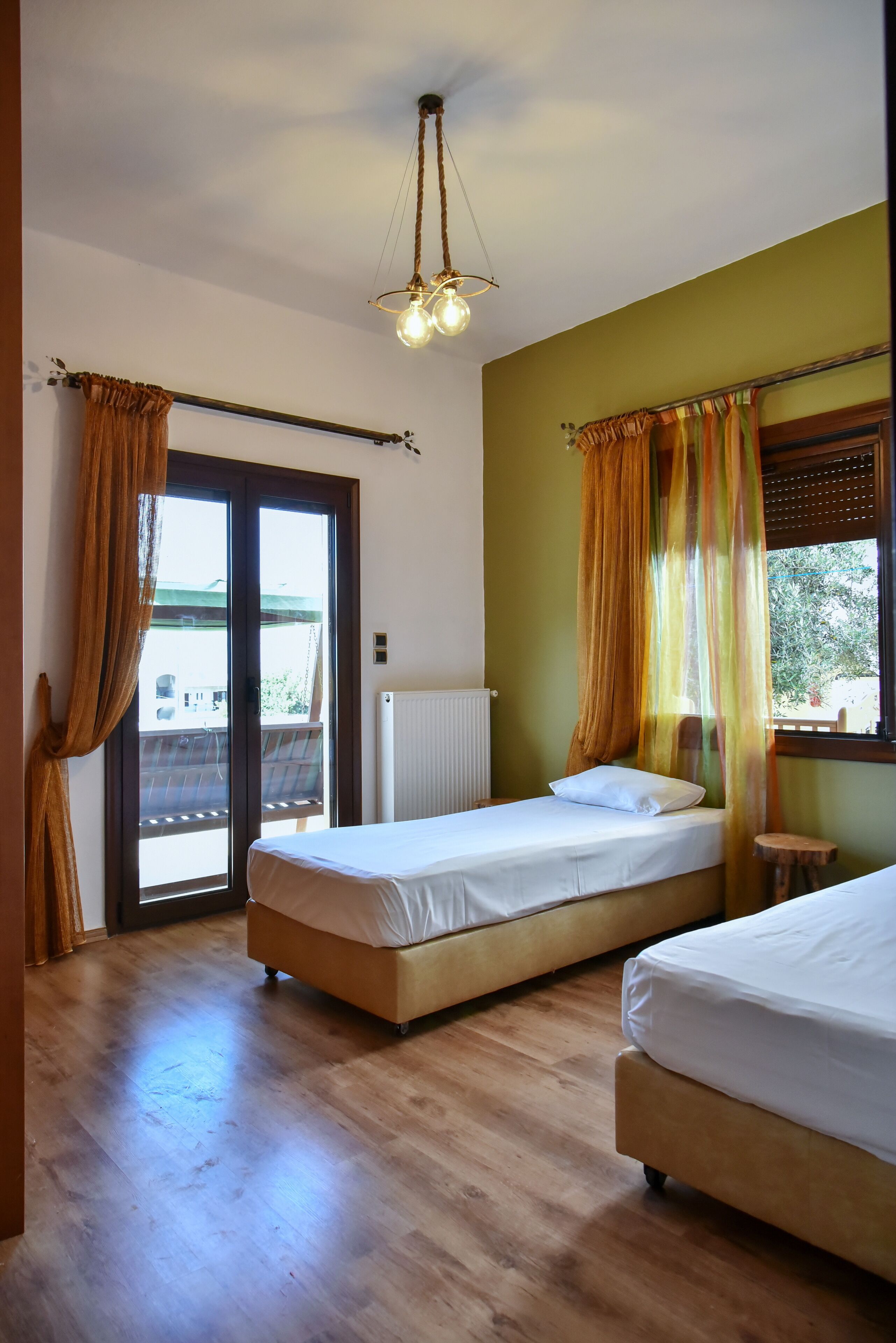 A room with two beds, a wooden floor, a ceiling light fixture, and a window with curtains.